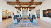 Located at 1230 Front Pl in North Port, Florida, this generously sized single-floor clubhouse is a showcase of contemporary design and thoughtful detail. The space is defined by dramatic soaring ceilings adorned with rich, dark wood beam accents that contrast beautifully against crisp white walls. Light herringbone-patterned hardwood flooring flows throughout, lending warmth and visual continuity across the entire layout.

The open living area is anchored by multiple seating vignettes, including plush sectional sofas, bold wingback accent chairs, and curated area rugs, all arranged to encourage both social gathering and relaxed lounging. A striking floor-to-ceiling slatted wood bookcase serves as an elegant room divider, adding architectural interest while preserving the open-concept feel. A built-in media wall flanked by custom shelving with textured blue-green backdrops and a sleek fireplace creates a refined focal point.

Oversized sliding glass doors and expansive multi-pane windows flood the interior with natural light while offering direct views of and access to an outdoor pool and cabana area. A sculptural starburst chandelier and a dramatic branching fixture add artistic flair overhead. Nature-inspired artwork, decorative wall installations, and curated accessories complete the sophisticated yet welcoming ambiance of this stylish community gathering space.