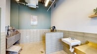 Located at 1624 W Babcock St in Bozeman, Montana, this single-room commercial space is thoughtfully designed as a dedicated pet grooming facility. The interior showcases a clean, modern aesthetic defined by subway tile wainscoting in muted sage and warm taupe tones that line the lower walls throughout. Polished concrete floors provide a durable, easy-to-maintain surface suited to the space's functional purpose.

The grooming area features a stainless steel wash tub with a step platform for easy pet access, complemented by a wall-mounted pet dryer and a utility sink. A tiled half-wall partition helps organize the workspace, while open wooden shelving displays decorative plants and grooming accessories, adding a welcoming touch. The accent wall in soft teal provides a calm, spa-like atmosphere.

The entry area is bright and open, framed by floor-to-ceiling windows and glass-panel doors that invite generous natural light and offer views of the tree-lined street outside. Exposed ductwork and industrial-style lighting overhead contribute to the contemporary commercial character of the space. A bike repair station near the entrance suggests a shared amenity context within a larger mixed-use building. Overall, the space balances practicality with a polished, inviting design.