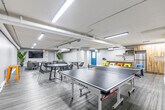 Located at 50 Glen Rd in Hamilton, Ontario, this amenity lounge is a well-appointed communal space designed for recreation and socializing. The open-concept layout is unified by durable wood-look vinyl flooring and a neutral grey palette throughout, creating a clean and modern atmosphere.

The space is divided into distinct functional zones. A games area anchors one side of the room, featuring a full-size ping-pong table set against a textured patterned accent wall. Adjacent to it is a kitchenette equipped with a sink, refrigerator, and microwave, complemented by a wooden island counter with colorful metal bar stools in yellow, orange, and green — adding a lively pop of color to the space.

A central dining and gathering area includes a round table with contemporary wire-frame chairs, ideal for casual meetings or group activities. On the lounge side, a pair of grey leather sofas create a comfortable seating arrangement, accompanied by a large wall-mounted flat-screen TV and a white media console, making it well-suited for movie nights or informal gatherings. Tall tropical plants add organic warmth to the corners of the room.

Structural elements such as exposed cylindrical support columns and overhead ductwork lend an industrial character to the space. Fluorescent strip lighting provides consistent, even illumination throughout. A teal-accented entry door with keypad access ensures secure entry, rounding out this fully functional amenity lounge.