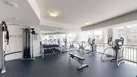 This fitness center offers a comprehensive workout environment housed within a single-level space designed with both functionality and comfort in mind. The layout is open and well-organized, with dark rubber flooring throughout providing durability and traction across all exercise zones.

The cardio area is a standout feature, lined with multiple treadmills and elliptical machines positioned directly in front of a continuous bank of large windows. These floor-to-ceiling windows flood the space with natural light and offer views of surrounding buildings and palm trees, creating an inviting atmosphere for extended workouts. A wall-mounted flat-screen television is also available in this zone.

The strength training area includes a well-stocked dumbbell rack with a full range of free weights, a flat weight bench, and a multi-functional cable machine suitable for a variety of resistance exercises. A leg press machine adds to the dedicated strength equipment selection. Full-length mirrors along one wall visually expand the space and support proper form during training.

The entrance area features double doors with windowed panels, a hand sanitizer dispenser, and a waste bin, emphasizing cleanliness and accessibility. Flush-mount ceiling lights provide consistent, even illumination throughout, complementing the natural light from the surrounding windows. The overall ambiance is clean, modern, and well-maintained.