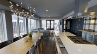 This contemporary single-floor space is defined by its open-concept layout and sophisticated design. The interior is anchored by a dramatic, open kitchen featuring a long white quartz waterfall island with a built-in gas cooktop, offering generous prep and seating space. Dark hardwood flooring runs throughout, creating a cohesive and elegant foundation for the entire space.

The dining area is outfitted with a natural wood table and modern gray chairs, adding warmth against the cool, dark gray ceiling and walls. A sculptural branching chandelier serves as a striking focal point overhead. The living area flows seamlessly from the dining space, furnished with a large sectional sofa and anchored by a plush area rug.

Floor-to-ceiling windows line the exterior walls, flooding the interior with abundant natural light and offering expansive views of the surrounding urban landscape. A set of glass-paned French doors opens directly to an outdoor terrace, blending interior and exterior living effortlessly. The overall ambiance is modern and refined, with a carefully curated palette of charcoal, white, and warm wood tones that lend the space a polished yet inviting character throughout.