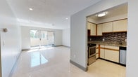 This single-floor, 2-bedroom, 1-bathroom unit offers a clean and functional layout designed to maximize space and natural light. The open living area is finished with large, high-gloss porcelain tile flooring that reflects light throughout the room, creating an airy and spacious ambiance. A textured ceiling adds architectural character overhead, while recessed lighting provides even illumination across the living space.

Sliding glass doors along the far wall flood the interior with natural light and provide direct access to a private outdoor area, framing views of surrounding greenery. The seamless transition between indoor and outdoor space enhances the overall sense of openness.

The kitchen is accessible through an arched entryway and features warm wood-toned cabinetry with cream upper cabinets, a dark granite countertop, and a mosaic tile backsplash that adds visual texture. Stainless steel appliances, including a range and refrigerator, lend a contemporary finish. Light tile flooring in the kitchen complements the overall neutral palette of the unit.

Together, the spaces form a cohesive, well-lit interior suited for comfortable everyday living.