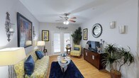 This single-floor residence offers a thoughtfully designed layout across several well-defined living spaces. The living room serves as the heart of the home, featuring warm hardwood-style flooring, a ceiling fan with light fixture, and French doors fitted with blinds that allow natural light to filter in. The neutral gray walls provide a versatile backdrop, while the open layout accommodates both a seating area and an entertainment space.

The kitchen is outfitted with warm-toned wood cabinetry extending to the ceiling, dark granite-style countertops, and black appliances including an electric range with microwave above. A double sink and ample storage make this a highly functional space. The adjacent dining area comfortably fits a full table and chairs, creating a natural flow between cooking and dining.

Two bedrooms each feature ceiling fans, carpeted floors, and windows with blinds and drapes that allow for natural light and privacy. Both are generously sized to accommodate full bedroom furnishings.

The two bathrooms mirror each other in finish quality, each offering granite-style countertops, wood cabinetry, large vanity mirrors with overhead lighting, and a tiled shower-tub combination with a black accent tile border.