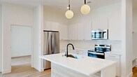 This thoughtfully designed single-floor residence offers a clean, contemporary aesthetic defined by crisp white walls, light wood-toned luxury vinyl flooring, and recessed lighting throughout. The open-concept kitchen serves as a central highlight, featuring a large waterfall-edge island with an undermount sink and matte black faucet, sleek white flat-panel cabinetry extending to the ceiling, stainless steel appliances including a French door refrigerator and electric range, and two globe pendant lights that add a modern, stylish touch.

The spacious living area opens directly to an outdoor balcony or patio via sliding glass doors, inviting natural light and providing a seamless indoor-outdoor connection. The bedroom is a comfortable, light-filled retreat with direct access to a walk-in closet outfitted with built-in shelving and hanging rods, offering ample organization. A second closet provides additional storage capacity.

All three bathrooms are finished to a high standard, featuring light wood vanities with white quartz countertops, large format mirrors, and marble-style porcelain tile surrounds. One bathroom includes a frameless glass walk-in shower, while another offers a full soaking tub-shower combination. The hallway connects all areas efficiently, contributing to the home's functional and well-organized flow.