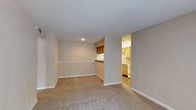 This single-floor residential space offers a clean, functional layout designed for comfortable everyday living. The main living area is finished with wall-to-wall carpet in a neutral beige tone, creating a warm and inviting atmosphere. Smooth, white ceilings with recessed lighting provide ample illumination, while the soft, neutral wall color throughout lends a cohesive and calming aesthetic. A defining architectural feature is the built-in pass-through counter with overhead cabinetry that connects the living area to the kitchen, adding both practicality and a sense of defined space without fully closing off the rooms. The kitchen, visible through an open doorway, features warm-toned wood cabinetry, white appliances, and hard surface flooring, offering a bright and efficient cooking space. The kitchen cabinetry extends to the ceiling, maximizing storage capacity. Clean white baseboards frame each room, adding a crisp finishing detail throughout. The overall layout flows naturally between the living and kitchen areas, making the space feel connected and well-organized. The understated, neutral design palette provides a versatile backdrop suitable for a variety of interior styles and furnishings.