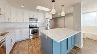 This single-story home offers a well-designed layout that balances open communal spaces with comfortable private retreats. The heart of the home is a striking open-concept kitchen and living area, where rich wood-look flooring flows seamlessly underfoot. The kitchen is outfitted with crisp white shaker cabinetry reaching to the ceiling, a classic subway tile backsplash, quartz countertops, and a full suite of stainless steel appliances. A standout blue-gray island with a white quartz surface anchors the space, complemented by a dramatic cluster of pendant lights overhead. A skylight adds additional natural brightness to the kitchen. The adjacent living area is bathed in natural light from large sliding glass doors that open to an outdoor balcony, with stylish pendant lighting enhancing the airy ambiance. The bedrooms feature wood-look flooring, ceiling fans with light fixtures, and generous closet space. The primary bathroom is a refined retreat, showcasing a classic black-and-white mosaic tile floor, a freestanding soaking tub, a separate glass-enclosed shower, dual vanity with dark countertops, and bold navy accent tile. A secondary bathroom offers a clean, modern look with white subway tile surrounding a tub-shower combination and a pedestal sink.
