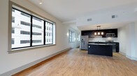 This single-floor space showcases a thoughtfully designed open-concept layout that seamlessly connects the living area and kitchen. Light wood-tone hardwood flooring runs throughout, lending warmth and continuity to the interior. Large, deep-set windows along the living area flood the space with natural light while framing views of the surrounding urban environment.

The kitchen is a standout feature, outfitted with dark espresso cabinetry that creates a bold contrast against the light walls and flooring. A kitchen island with a dark countertop serves as both a functional workspace and a visual anchor for the room. Stainless steel appliances, including a French door refrigerator, complement the modern aesthetic. Pendant lighting with warm-toned bulbs hangs above the island, adding an inviting ambiance to the cooking and dining area.

The living space is open and generously proportioned, offering flexibility for various furniture arrangements. Recessed lighting in the ceiling supplements the natural light from the windows, ensuring the space remains bright throughout the day. The clean, minimalist design with neutral white walls and crisp trim creates a versatile backdrop suited for a range of interior styles. Overall, the space exudes a contemporary urban character with refined, functional details.