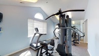 This dedicated fitness room offers a comprehensive workout environment designed for convenience and functionality. The space is characterized by a vaulted ceiling that lends an open, airy feel, while a charming arched window allows natural light to fill the room, complementing the soft blue walls and light-toned flooring. A large multi-station cable machine — a Body-Solid unit — serves as the centerpiece of the room, providing a wide range of strength training options. A treadmill with an integrated display console sits nearby, catering to cardio exercise needs. Additional equipment is visible reflected in a wall-mounted mirror, which also serves to visually expand the space and allows users to monitor their form during workouts. Horizontal blinds on the windows offer privacy and light control as needed. The flooring appears to be a durable, textured surface well-suited for a fitness environment. Overall, the room is thoughtfully arranged to maximize usability within a compact footprint, making it a practical and inviting space for a complete daily workout routine.
