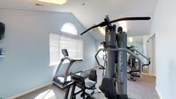 This dedicated fitness room offers a comprehensive workout environment designed for convenience and functionality. The space is characterized by a vaulted ceiling that lends an open, airy feel, while a charming arched window allows natural light to fill the room, complementing the soft blue walls and light-toned flooring. A large multi-station cable machine — a Body-Solid unit — serves as the centerpiece of the room, providing a wide range of strength training options. A treadmill with an integrated display console sits nearby, catering to cardio exercise needs. Additional equipment is visible reflected in a wall-mounted mirror, which also serves to visually expand the space and allows users to monitor their form during workouts. Horizontal blinds on the windows offer privacy and light control as needed. The flooring appears to be a durable, textured surface well-suited for a fitness environment. Overall, the room is thoughtfully arranged to maximize usability within a compact footprint, making it a practical and inviting space for a complete daily workout routine.