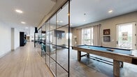Located at 383-389 Main St, this generously sized single-floor amenity lounge offers a thoughtfully designed communal space ideal for socializing and recreation. The open-plan layout spans a substantial footprint, with distinct zones flowing seamlessly into one another via wide-plank wood-look flooring that runs throughout most of the space.

At one end, a stylish wet bar with a dark cabinetry island, bar stools, and pendant lighting anchors a kitchen-style gathering area, complemented by large windows and glass door access that fills the space with natural light and views of the surrounding streetscape. Moving through the space, multiple seating arrangements — including sofas, accent chairs in teal, and high-top tables — create versatile lounge zones, accented by bold teal feature walls, open shelving displays, and curated artwork.

A floor-to-ceiling steel-framed glass partition wall elegantly separates the billiards area, which features a full-sized pool table set atop patterned carpet, wall-mounted cue storage, and large draped windows overlooking an outdoor terrace. Recessed lighting throughout ensures a well-lit environment across all zones. The overall ambiance is contemporary and inviting, blending industrial design accents with warm tones to create a dynamic, multi-use community retreat.