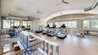 This two-level fitness center in Phoenix, AZ offers a generously sized, thoughtfully designed workout facility spread across two exercise floors. The main floor greets visitors with striking herringbone-patterned wood flooring, textured 3D white wall panels, and full-length mirrors that amplify the sense of space and light. This level is equipped with a comprehensive free weights area featuring dumbbells, kettlebells, medicine balls, and barbells, alongside cable machines, weight benches, and a squat rack. A dedicated cycling studio with Spinning bikes and floor-to-ceiling mirrors provides a focused space for indoor cycling workouts. A sleek metal staircase, accented by a bold, blue patterned mosaic accent wall, connects to the upper level. The second floor features soaring vaulted ceilings with exposed ductwork and large windows on multiple sides, flooding the space with natural light and offering views of the surrounding greenery. This level houses a row of treadmills, elliptical machines, and stationary bikes, complemented by ceiling fans for added comfort. Throughout both floors, the design balances modern aesthetics with functional fitness infrastructure, creating an energetic yet inviting environment.