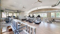 Located at 1550 East Campbell Avenue in Phoenix, AZ, this two-floor fitness center offers a comprehensive and well-appointed exercise facility designed for a full range of workout styles. The larger main level provides generous open space with a mix of polished concrete and striking herringbone-patterned hardwood flooring. Soaring double-height ceilings with exposed ductwork and large multi-pane windows flood the floor with natural light and an open, airy ambiance. This level houses a robust free weights area stocked with dumbbells, kettlebells, medicine balls, and barbells, alongside cable machines, weight benches, and a squat rack. A dedicated spin studio with Spinning bikes and floor-to-ceiling mirrors adds a boutique fitness feel. Three-dimensional textured white wall panels and a bold blue-patterned accent wall provide distinctive visual character throughout. The upper level, accessed by a sleek open staircase with cable railings, features vaulted ceilings, ceiling fans, and panoramic windows overlooking lush outdoor greenery. This floor is equipped with a row of treadmills, ellipticals, and stationary bikes, alongside storage for yoga mats and accessories. Full-length mirrors amplify the sense of space on both levels, creating a bright, motivating environment suited for any fitness regimen.