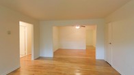 This single-floor space presents a clean and functional layout defined by warm hardwood flooring that runs throughout, creating a sense of continuity and cohesion across the interior. The living areas are openly connected through a wide architectural passageway, allowing for a natural flow between spaces. This open-concept design enhances the overall sense of roominess and flexibility in how the space can be arranged and utilized.

The interior features neutral white walls that provide a bright, airy backdrop well-suited to a variety of décor styles. A ceiling fan is installed in the adjoining room, contributing both to comfort and to the practical character of the space. White baseboards and trim add a crisp finishing detail throughout.

The hardwood floors, with their warm honey tones, are a standout feature, lending both durability and timeless appeal. The overall ambiance is understated and versatile — an inviting blank canvas ready to be personalized. The open passage between rooms encourages an easy, interconnected living experience, making the space feel expansive and well-proportioned for everyday living.