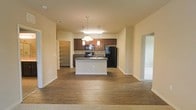 This single-floor space is designed with an open-concept layout that promotes a sense of spaciousness and connectivity between living areas. The kitchen serves as the focal point, anchored by a central island with a built-in sink and dark countertop, providing both a functional workspace and a natural gathering point. Rich, dark wood cabinetry lines the kitchen walls, complemented by black appliances including a refrigerator and range, creating a cohesive and contemporary aesthetic. Pendant lighting hangs above the island, offering focused task lighting while adding a decorative element to the space.

The flooring transitions from hardwood-style planks in the kitchen and main living area to carpeting in the adjacent room, distinguishing different zones within the open floor plan. A walk-in pantry or storage closet is accessible just off the kitchen, providing convenient organizational space. Neutral, warm-toned walls and standard ceiling heights contribute to a clean, understated interior throughout. Doorways lead to additional rooms, suggesting a well-connected and functional single-level floor plan suited for comfortable everyday living.