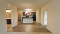 This single-floor space is designed with an open-concept layout that promotes a sense of spaciousness and connectivity between living areas. The kitchen serves as the focal point, anchored by a central island with a built-in sink and dark countertop, providing both a functional workspace and a natural gathering point. Rich, dark wood cabinetry lines the kitchen walls, complemented by black appliances including a refrigerator and range, creating a cohesive and contemporary aesthetic. Pendant lighting hangs above the island, offering focused task lighting while adding a decorative element to the space.

The flooring transitions from hardwood-style planks in the kitchen and main living area to carpeting in the adjacent room, distinguishing different zones within the open floor plan. A walk-in pantry or storage closet is accessible just off the kitchen, providing convenient organizational space. Neutral, warm-toned walls and standard ceiling heights contribute to a clean, understated interior throughout. Doorways lead to additional rooms, suggesting a well-connected and functional single-level floor plan suited for comfortable everyday living.