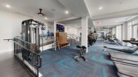 Located at 3011 Hidden Forest Ct in Marietta, GA, this professionally designed fitness facility spans a generous single floor and is divided into two distinct exercise rooms, each thoughtfully appointed for a comprehensive workout experience.

The main exercise room is an expansive, open-plan space featuring large floor-to-ceiling windows that flood the interior with natural light and offer views of the surrounding greenery. Vibrant, patterned carpet tiles in shades of blue and gray line the floor, while ceiling fans provide comfortable air circulation throughout. The room is equipped with a full array of cardio machines—including treadmills, ellipticals, and rowing equipment—alongside multi-station strength-training systems, free weights, and functional training accessories. Motivational athletic murals and bold directional graphics energize the walls, and a wall-mounted flat-screen TV adds to the amenity-rich environment. Wooden lockers provide convenient storage near the entry staircase.

The second exercise room offers a studio-style setting, finished with smooth flooring, a full-length mirrored wall with a ballet barre, and a wall-mounted TV. Stationary cycling bikes line one side, making it ideal for group fitness or individual training sessions. A contemporary linear pendant light fixture and recessed lighting create a polished, focused atmosphere. A glass partition wall connects both spaces visually, maintaining an open, cohesive feel throughout the facility.
