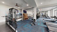 Located at 3011 Hidden Forest Ct in Marietta, GA, this professionally designed fitness facility spans a generous single floor and is divided into two distinct exercise rooms, each thoughtfully appointed for a comprehensive workout experience.

The main exercise room is an expansive, open-plan space featuring large floor-to-ceiling windows that flood the interior with natural light and offer views of the surrounding greenery. Vibrant, patterned carpet tiles in shades of blue and gray line the floor, while ceiling fans provide comfortable air circulation throughout. The room is equipped with a full array of cardio machines—including treadmills, ellipticals, and rowing equipment—alongside multi-station strength-training systems, free weights, and functional training accessories. Motivational athletic murals and bold directional graphics energize the walls, and a wall-mounted flat-screen TV adds to the amenity-rich environment. Wooden lockers provide convenient storage near the entry staircase.

The second exercise room offers a studio-style setting, finished with smooth flooring, a full-length mirrored wall with a ballet barre, and a wall-mounted TV. Stationary cycling bikes line one side, making it ideal for group fitness or individual training sessions. A contemporary linear pendant light fixture and recessed lighting create a polished, focused atmosphere. A glass partition wall connects both spaces visually, maintaining an open, cohesive feel throughout the facility.
