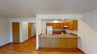 This single-floor unit is thoughtfully laid out to maximize functionality across its living spaces. The kitchen serves as a central highlight, featuring warm honey-toned oak cabinetry extending to the ceiling for ample storage, granite-look countertops, and white appliances including a Whirlpool electric range with an overhead ventilation hood and a full-size refrigerator. A peninsula with matching countertop and oak paneling separates the kitchen from the adjacent living area, providing a practical breakfast bar while maintaining an open, connected feel. Recessed lighting illuminates the kitchen workspace effectively.

The living areas are finished with light-toned hardwood flooring, which extends through the main living space and into at least one bedroom. A sliding glass door fitted with vertical blinds allows natural light to flow into the living room and provides outdoor access. A secondary bedroom features carpeting and a window overlooking the exterior. The bathroom is equipped with a full tub-shower combination in white, complemented by tile flooring. Throughout the unit, textured ceilings, neutral wall tones, and consistent wood-toned trim elements create a cohesive and practical interior aesthetic.