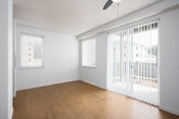 This single-floor unit is designed with a clean, contemporary aesthetic throughout. The living area is an open, airy space highlighted by warm light wood flooring, crisp white walls, and a ceiling fan overhead. Natural light flows in generously through a large sliding glass door fitted with vertical blinds, offering direct access to an outdoor area and views of surrounding greenery. An additional side window further enhances the bright, welcoming atmosphere.

The bedroom is a well-proportioned room featuring the same light wood flooring and white walls, with two windows dressed in horizontal blinds that allow for ample natural light. A door adjacent to the bedroom provides convenient access to the rest of the unit.

The kitchen is a functional and stylish space outfitted with two-tone cabinetry — slate blue lower cabinets paired with light upper cabinets — complemented by clean white countertops and a subtle light blue backsplash. Stainless steel appliances, including a range, over-the-range microwave, and refrigerator, complete the modern look.

The bathroom features a straightforward, clean design with white walls, a bathtub, and brushed nickel hardware including grab bars and towel rails. The unit's consistent use of light tones and wood-style flooring throughout creates a cohesive and inviting living environment.
