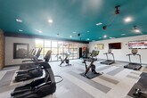 This modern fitness center offers a generously sized, single-floor exercise space designed for a comprehensive workout experience. The room's most striking architectural feature is its vibrant teal ceiling, which adds a bold, energizing aesthetic complemented by recessed lighting throughout. A striking floor-to-ceiling glass wall runs along one side of the space, flooding the interior with natural light and providing direct views of an outdoor pool and patio area, creating a seamless connection between indoor fitness and outdoor amenity spaces. The flooring features a patterned, multi-tone vinyl tile in varying shades of gray, lending a clean and contemporary look to the space. One wall showcases a warm wood-panel accent, adding texture and visual contrast to the otherwise neutral palette. A full-length mirror along one wall enhances the sense of depth and openness. The fitness center is well-stocked with a variety of cardio equipment, including treadmills, cycling bikes, and elliptical machines, as well as strength training options such as a dumbbell rack and a cable machine. Multiple wall-mounted flat-screen TVs are positioned throughout for entertainment during workouts. Exercise balls and accessories are neatly stored along one wall, rounding out the facility's comprehensive fitness offerings.