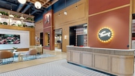 Located at 415 N Lake St in Madison, WI, this dynamic single-floor commercial space spans 4,780 square feet of thoughtfully designed interior. The open-plan layout features soaring ceilings with exposed industrial ductwork painted in slate blue, complemented by warm wood coffered ceiling panels and an eye-catching array of globe pendant lights in varying sizes and finishes, including a striking gold cluster chandelier. Polished concrete floors flow throughout, punctuated by bold patterned tile inlays and colorful area rugs that define distinct zones. The space is anchored by a central reception desk bearing a neon "Theory" sign, set against a deep terracotta accent wall with plaid wallcovering. A central banquette seating island with a marble-top bar counter and teal stools provides a casual dining hub, while open shelving units display lush greenery and books overhead. Floor-to-ceiling windows along the exterior walls flood the space with natural light and offer outdoor views. Additional seating zones include lounge areas with eclectic upholstered furniture, private booth seating visible behind glass partitions, and a counter bar with bar-height stools. Playful branding, vibrant artwork, and curated plant installations complete the lively, welcoming ambiance throughout.