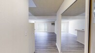 This single-floor unit offers a well-organized layout comprising a living room, kitchen, and bathroom. The living room serves as the central space, finished with light wood-look laminate flooring and neutral beige walls that lend a clean, understated aesthetic. A large window fitted with vertical blinds allows generous natural light to fill the room, while a wall-mounted air conditioning unit provides climate control. The textured ceiling adds a subtle visual layer to the open feel of the room. The kitchen is efficiently designed with a peninsula-style layout featuring a tiled countertop and darker granite-look work surface. White upper cabinetry provides ample storage, and the space is equipped with a white gas range, a full-size refrigerator, and an exhaust hood. The light wood flooring continues seamlessly from the living area into the kitchen, reinforcing the cohesive design. A mirrored sliding closet is visible near the kitchen entry, offering additional storage. The bathroom is straightforward and functional, featuring a full-size tub and shower combo with chrome fixtures, a toilet, and a towel ring — all finished in a clean white palette with light flooring. Throughout the unit, the consistent use of light tones and wood-look flooring creates a unified and easy-to-maintain living environment.