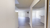 This single-floor unit offers a well-organized layout comprising a living room, kitchen, and bathroom. The living room serves as the central space, finished with light wood-look laminate flooring and neutral beige walls that lend a clean, understated aesthetic. A large window fitted with vertical blinds allows generous natural light to fill the room, while a wall-mounted air conditioning unit provides climate control. The textured ceiling adds a subtle visual layer to the open feel of the room. The kitchen is efficiently designed with a peninsula-style layout featuring a tiled countertop and darker granite-look work surface. White upper cabinetry provides ample storage, and the space is equipped with a white gas range, a full-size refrigerator, and an exhaust hood. The light wood flooring continues seamlessly from the living area into the kitchen, reinforcing the cohesive design. A mirrored sliding closet is visible near the kitchen entry, offering additional storage. The bathroom is straightforward and functional, featuring a full-size tub and shower combo with chrome fixtures, a toilet, and a towel ring — all finished in a clean white palette with light flooring. Throughout the unit, the consistent use of light tones and wood-look flooring creates a unified and easy-to-maintain living environment.