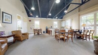 Located at 7200 Gorden Farms Parkway in Dublin, Ohio, this single-floor gathering space offers a welcoming and functional environment designed for community use. The room's most striking architectural feature is its dramatic vaulted ceiling, finished in deep navy blue and accented by exposed dark structural beams that run the length of the space. Pendant lights and track lighting are suspended from the ceiling, providing warm, even illumination throughout.

The open layout is divided into distinct functional zones. A comfortable lounge area near the entrance features plush leather armchairs and a sofa arrangement, complemented by framed artwork and a side table with a lamp. Multiple dining and meeting areas are furnished with classic wooden tables and chairs, providing ample seating for gatherings or events.

Large multi-pane windows and a set of French doors flood the interior with natural light and offer views of the surrounding greenery. The neutral, warm beige walls and soft carpeted floors enhance the inviting ambiance throughout the main area.

A dedicated kitchenette along one wall is equipped with white shaker-style cabinetry, granite countertops, a built-in microwave, and stone tile flooring—ideal for catering or refreshment service. A water cooler and coffee station are also conveniently accessible. The space combines practical amenities with a charming architectural character.