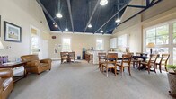 Located at 7200 Gorden Farms Parkway in Dublin, Ohio, this single-floor gathering space offers a welcoming and functional environment designed for community use. The room's most striking architectural feature is its dramatic vaulted ceiling, finished in deep navy blue and accented by exposed dark structural beams that run the length of the space. Pendant lights and track lighting are suspended from the ceiling, providing warm, even illumination throughout.

The open layout is divided into distinct functional zones. A comfortable lounge area near the entrance features plush leather armchairs and a sofa arrangement, complemented by framed artwork and a side table with a lamp. Multiple dining and meeting areas are furnished with classic wooden tables and chairs, providing ample seating for gatherings or events.

Large multi-pane windows and a set of French doors flood the interior with natural light and offer views of the surrounding greenery. The neutral, warm beige walls and soft carpeted floors enhance the inviting ambiance throughout the main area.

A dedicated kitchenette along one wall is equipped with white shaker-style cabinetry, granite countertops, a built-in microwave, and stone tile flooring—ideal for catering or refreshment service. A water cooler and coffee station are also conveniently accessible. The space combines practical amenities with a charming architectural character.