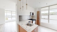 This contemporary two-floor space showcases a clean, minimalist aesthetic throughout, characterized by crisp white walls, light wood-tone flooring, and an abundance of natural light. The open-plan kitchen is well-appointed with stainless steel appliances including a gas range, built-in microwave, and dishwasher, complemented by sleek white upper cabinetry, warm wood-toned lower cabinetry, a stainless steel backsplash, and a peninsula countertop with an undermount sink. Pendant lighting above the peninsula adds a refined finishing touch. The upper level features a bright, airy living area framed by floor-to-ceiling windows on two sides, offering sweeping panoramic views of the surrounding urban landscape and rolling hills beyond. Light pours generously into every corner, enhancing the open and airy atmosphere. A staircase with a modern stainless steel horizontal-rail railing connects the two levels, providing a stylish architectural detail. The bathroom is finished with a vessel sink set atop a floating wood vanity, a large mirror with integrated storage, and clean white surfaces throughout. The overall ambiance is bright, modern, and thoughtfully designed, with consistent use of warm wood accents balanced against white surfaces to create a welcoming yet contemporary feel.