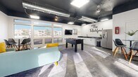 Located at 1207 E 8th St in Tempe, Arizona, this community lounge space for The Rockford offers a thoughtfully designed, single-floor open-plan layout that blends modern industrial aesthetics with comfortable social amenities. The space is defined by its striking contrast of crisp white walls against a dramatic matte-black ceiling, through which exposed HVAC ductwork and flush panel lighting run throughout, lending a contemporary, urban character to the room.

Polished concrete floors extend across the entire space, providing a durable and visually cohesive foundation. Large banks of white-framed windows line the perimeter walls, flooding the interior with generous natural light and offering open views of the surrounding Tempe neighborhood. French doors provide direct access to an outdoor area, further enhancing the sense of connectivity to the exterior.

The lounge is furnished with a sectional sofa and accent seating arranged around area rugs, creating a relaxed gathering zone. A foosball table serves as a central recreational focal point, complemented by a wall-mounted flat-screen television. A bistro-style dining table with modern chairs offers an additional casual seating option. A kitchenette along one wall features white cabinetry, a dark countertop, a sink, and a stainless steel refrigerator, making the space well-equipped for social events. A closet provides convenient storage. The Rockford branding is prominently displayed on the wall, completing the space's cohesive identity.