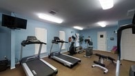 This fitness room offers a practical and functional workout environment designed to support a range of exercise routines. The space is finished with light blue walls and a durable cork-style flooring, creating a clean and comfortable atmosphere for physical activity. Overhead fluorescent lighting ensures the room is well-lit throughout, supplemented by natural light streaming in through multiple windows fitted with white horizontal blinds.

The room is equipped with several pieces of cardio equipment, including two Spirit-brand treadmills positioned along the windowed wall, as well as an elliptical machine for low-impact training. A stationary recumbent-style bike and an upright rowing or resistance machine round out the cardio offerings. A weight bench with attachments is also present, providing options for strength and resistance training.

A decorative potted plant adds a touch of greenery to the space, while a fire extinguisher is mounted on the wall, reflecting attention to safety. A digital access panel is visible near the exit door, suggesting controlled entry. The room's single-floor layout and straightforward design make it an accessible and efficient facility for everyday fitness use.
