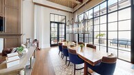 This well-appointed clubhouse in Royal Palm Beach, Florida offers a thoughtfully designed single-floor space ideal for community gatherings and events. The layout is anchored by a dramatic dining area showcasing soaring ceilings with exposed wood beams, floor-to-ceiling steel-framed windows, and French doors that flood the room with natural light and provide views of a lush outdoor patio. A large live-edge wood dining table is paired with rich navy and leather upholstered chairs, while a row of elegant glass pendant chandeliers adds a refined, warm ambiance overhead. Decorative patterned tile accents the hardwood flooring throughout, adding a distinctive visual touch. Adjacent to the dining space is a fully equipped kitchen featuring warm wood cabinetry with glass-front upper panels, stainless steel appliances including a French door refrigerator, double wall ovens, a professional range hood, and a striking blue-and-white herringbone tile backsplash. A central island with quartz countertops provides additional prep and seating space. The clubhouse also includes a private screening room with high ceilings accented by a coffered wood ceiling detail, cool grey walls, plush leather lounge chairs arranged around a central table, and a wall-mounted projection screen — creating an intimate, cinema-like retreat.