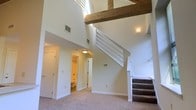 This two-story residence makes a striking impression with its dramatic double-height ceiling in the main living area, which draws the eye upward and creates an expansive sense of openness. A prominent exposed wooden beam runs overhead, adding a warm, rustic architectural detail that contrasts beautifully with the clean white walls throughout the space. The living area is finished with wall-to-wall carpeting, contributing to a comfortable and cohesive feel underfoot. Natural light enters from upper-level windows, brightening the interior and highlighting the airy vertical proportions of the main room. A carpeted staircase with a white railing leads to the second floor, offering a clean and functional transition between levels. On the main floor, a bathroom is conveniently accessible, featuring standard fixtures and warm lighting. Ample wall outlets and HVAC vents are positioned thoughtfully throughout, ensuring comfort and practicality. The neutral color palette of soft whites and warm beiges lends the space a calm, versatile ambiance well-suited to a variety of furnishing styles. Overall, the combination of high ceilings, exposed timber, and an open floor plan gives this residence a distinctive character that balances modern simplicity with rustic warmth.