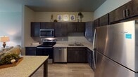 This single-floor residence at 2016 Main Ave S in Brookings, South Dakota, offers a thoughtfully designed layout that balances functionality with modern style. The kitchen serves as a centerpiece, outfitted with dark espresso cabinetry, granite-style countertops, and a full suite of stainless steel appliances including a refrigerator, electric range, over-the-range microwave, and dishwasher. Light wood-tone flooring runs throughout the kitchen and connects seamlessly to the dining area, where a granite-topped island with seating provides additional workspace and casual dining space. The living area is open and inviting, featuring neutral gray walls and a large sliding glass door with vertical blinds that allows natural light and outdoor views. The bedroom offers a comfortable, carpeted retreat with a window allowing natural light. A hallway connects the bedroom to the bathroom, which features dark vanity cabinetry with a granite-style countertop, a framed mirror, wood-look flooring, and a shower with a curtain rod. Throughout the unit, consistent neutral tones, modern fixtures, and cohesive finishes create a clean and contemporary ambiance.