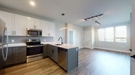 This thoughtfully designed single-floor residence showcases a contemporary aesthetic with a seamless blend of style and function. The open-concept kitchen is a standout feature, boasting two-tone cabinetry — crisp white upper cabinets paired with slate-blue lower cabinets — topped with clean white quartz countertops. A peninsula island with an undermount sink and matte black faucet serves as a natural focal point, complemented by a full suite of stainless steel appliances including a refrigerator, range, microwave, and dishwasher. A linear pendant light adds a modern touch overhead, while wood-look flooring flows throughout the main living areas, creating warmth and continuity.

The living area opens naturally from the kitchen, offering a bright and welcoming space bathed in natural light from a large window. Two bedrooms are finished with plush neutral carpeting and generous windows that frame lush outdoor views, filling each room with an abundance of natural light.

The bathrooms reflect the same modern sensibility found throughout, featuring wood-look tile flooring, white floating or wall-mounted vanities with matte black hardware, and clean, minimal design. The overall palette of soft grays, whites, and warm wood tones creates a cohesive, move-in-ready environment that is both functional and visually refined.