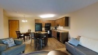 This single-floor space offers an open-concept layout that seamlessly connects the kitchen, dining area, and living room, creating a cohesive and functional living environment. The kitchen is well-appointed with warm medium-toned wooden cabinetry, granite-look countertops, and a full suite of black appliances including a refrigerator, stove, microwave, and dishwasher. A central kitchen island with bar seating provides additional counter space and a casual dining option. The flooring throughout the main living area is durable hardwood-style planking in a rich, warm tone that complements the cabinetry. The dining area accommodates a full table and chair set, illuminated by an elegant chandelier that adds a touch of classic charm. The living room flows naturally from the dining and kitchen spaces, offering a comfortable seating arrangement. Overhead lighting keeps the space bright and welcoming. The warm neutral wall tones throughout enhance the inviting ambiance, while the open floor plan ensures excellent sightlines and a sense of spaciousness across the entire main living area.