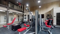 Located at 321 E Capital St in Columbus, Ohio, this fitness center delivers a well-appointed, professional-grade workout facility on a single floor. The space is entered through sleek, floor-to-ceiling glass doors that open onto polished concrete flooring in the entryway, setting a modern and industrial tone. Inside the main workout area, patterned blue-gray carpet tiles cover the floor beneath a collection of Matrix-brand strength and cardio equipment, including cable machines, adjustable benches, leg press stations, and a variety of free weights on a dedicated dumbbell rack. Cardio equipment includes treadmills, ellipticals, and a stair-climber, arranged to allow ample circulation throughout the space. Soaring ceilings with clerestory windows along the upper walls invite natural light deep into the room, while recessed and pendant lighting provide consistent illumination. Textured concrete-finish walls add an urban, loft-inspired aesthetic that contrasts with the crisp white trim and painted walls. Full-length mirrors are strategically placed to maximize sightlines and enhance the sense of spaciousness. A wall-mounted flat-screen television is positioned for visibility across the main workout zone. Plyometric boxes and floor mats round out the functional training offerings, making this facility suitable for a wide range of fitness routines.
