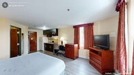 Located at 1340 N Wesleyan Blvd in Rocky Mount, NC, this self-contained studio suite offers a thoughtfully arranged single-floor layout designed for both comfort and functionality. The main living area features light wood-style flooring, warm neutral wall tones, and an accent wall behind the king-size bed, creating a welcoming ambiance. A window dressed with rich burgundy curtains allows natural light to fill the room, complemented by bedside lamps for a cozy atmosphere. The sleeping area is furnished with a large bed, a wood-tone dresser, and a flat-screen TV, providing all essential amenities within easy reach. A dedicated workspace includes a desk and ergonomic chair, ideal for extended stays. The integrated kitchenette is outfitted with dark cabinetry, a refrigerator, microwave, stovetop, and sink with tile backsplash — offering convenient in-room meal preparation. A wall-mounted ironing board near the kitchenette adds practical utility. The bathroom features tile-style flooring, a vanity with a dark granite countertop, a wall mirror, and a combined shower/tub, finished in soft yellow-green tones. The suite's efficient design maximizes every square foot, making it a well-equipped space for short or extended stays.