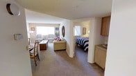 This single-floor residence is thoughtfully laid out to maximize comfort and functionality. The living area serves as the heart of the home, decorated in a charming nautical theme with coastal accents including an anchor wall hanging and a decorative rope mirror. Plush carpeting in warm neutral tones runs throughout, lending a soft, welcoming feel underfoot. A large window floods the living space with natural light, illuminating a seating arrangement that includes a cushioned sofa with accent pillows, two classic accent chairs, and a rustic storage trunk that doubles as a coffee table. A dining nook is positioned just off the main corridor, featuring a round bistro-style table with wooden chairs — an ideal spot for casual meals. Adjacent to the living area, the bedroom is accessible through a wide doorway and features its own window with sheer curtains, a full-size bed dressed in bold navy and white striped bedding, and a cozy, restful ambiance. Warm oak cabinetry is visible nearby, suggesting additional storage or a kitchenette area. The textured ceilings and neutral wall tones throughout create a consistent, understated backdrop that ties each area of the space together cohesively.