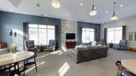 This single-floor lounge space is designed with both comfort and style in mind. The room's most striking feature is the floor-to-ceiling stacked stone accent wall, which serves as a focal point and anchors a wall-mounted flat-screen television atop a rich, red-toned media console. Deep teal-painted walls contrast elegantly with the neutral stone, creating a bold yet inviting color palette throughout.

The seating area is generously sized, accommodating a large grey leather sectional sofa paired with patterned armchairs in blue and white tones, offering a variety of comfortable seating options. Pendant and flush-mount light fixtures hang from the tray ceiling, complementing the recessed lighting that illuminates the space evenly.

Natural light pours in through multiple large windows dressed with dark curtain panels, framing views of the surrounding exterior. The soft, neutral carpeting adds warmth underfoot and contributes to the room's relaxed ambiance. Overall, the space balances modern design elements with comfortable furnishings, making it well-suited as a communal gathering or entertainment area.