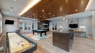 Located at 2599 Evans Rd in Morrisville, NC, this single-floor community amenity space is thoughtfully designed to encourage gathering, recreation, and relaxation. The open-concept layout flows seamlessly between distinct zones, all unified by warm hardwood-style flooring and a striking slatted wood ceiling with recessed and ambient lighting that adds depth and warmth throughout.

The main game and lounge area is a standout, featuring a pool table, shuffleboard, and a large dark island with bar-height seating illuminated by sculptural pendant fixtures. Generous sectional sofas, layered rugs, and accent chairs create an inviting lounge zone anchored by a wall-mounted flat-screen TV. Large windows along multiple walls flood the space with natural light and offer views of the surrounding community.

A dedicated kitchen area serves the space with practical elegance. Gray shaker-style cabinetry pairs with light stone countertops, while a bold Arabesque-patterned tile backsplash in teal and white adds a decorative focal point. Open floating shelves in dark wood display serveware and accessories, completing the functional yet stylish design. The overall ambiance is modern and sophisticated, balancing comfort with a refined, upscale aesthetic ideal for community events and everyday leisure.