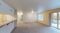 This single-floor unit features a well-thought-out layout connecting key living spaces through a central hallway. The spacious living area is finished with neutral-toned carpet and warm cream-colored walls, creating a bright and inviting atmosphere. A large window with horizontal blinds allows natural light to fill the room, with views of surrounding greenery. A wall-mounted heating vent ensures comfortable climate control. An adjacent sliding door provides additional outdoor access.

The galley-style kitchen is efficiently designed with light wood cabinetry, speckled countertops, and wood-look vinyl flooring. An electric range and sink are neatly positioned along opposing walls, maximizing workspace. Notably, the kitchen opens directly to an in-unit laundry area, housing a full-sized washer and dryer — a practical and convenient feature.

The bedrooms are carpeted and well-proportioned, each featuring large windows with blinds that frame outdoor views of trees and surrounding grounds. One bedroom includes a sliding panel closet door, offering organized storage.

The bathrooms are consistent in design, each featuring a tub and shower combination enclosed by frosted sliding glass doors, a wood-toned vanity with a speckled countertop, a single sink, and wood-look vinyl flooring throughout.
