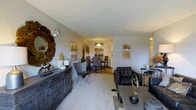 This single-floor space is designed with an open-concept layout that seamlessly connects the living, dining, and kitchen areas, creating a sense of spaciousness and flow throughout. The living area is carpeted and furnished with dark leather seating, a glass-top coffee table, and an ornate wooden sideboard adorned with decorative accents, including a sculptural mirror and wall art in warm, earthy tones. The neutral-toned walls provide a versatile backdrop that complements both the furnishings and décor.

Moving toward the dining area, the flooring transitions from carpet to a harder surface, adding visual distinction between the zones. A pendant light fixture hangs above the dining table, which is surrounded by wooden chairs, creating an inviting setting for meals. Beyond the dining area, a glimpse of the kitchen reveals white cabinetry and ample counter space, suggesting a functional and bright cooking environment. The overall ambiance of the space is warm and welcoming, with consistent neutral tones, layered textures, and carefully chosen décor contributing to a cohesive and comfortable interior aesthetic.