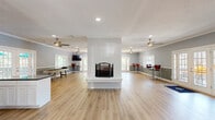 Located at 100 Pinegate Cir in Chapel Hill, NC, this well-appointed single-floor space offers a generous, open-concept layout designed for gathering and community use. Light hardwood flooring flows throughout, complementing the cool blue-gray walls, crown molding, and textured ceilings that define the interior's clean, welcoming aesthetic.

The kitchen is a functional highlight, featuring white shaker cabinetry, dark granite countertops, a central island, subway tile backsplash, and a full suite of stainless steel appliances including a refrigerator, range, and built-in microwave. The kitchen opens seamlessly into the expansive living area, creating an ideal environment for social interaction.

The living room is generously proportioned, anchored by a striking white-surround fireplace that serves as a central focal point. Dual ceiling fans with recessed lighting keep the space comfortable, while multiple sets of French doors and large windows invite abundant natural light and provide views of the surrounding greenery. A dedicated seating area with leather furniture and a high-top bar area with stools offer versatile zones for relaxation or casual dining.

A hallway connects the main areas, and the layout as a whole is practical and easy to navigate. The overall ambiance is bright, modern, and inviting — well-suited for community events, meetings, or everyday resident use.