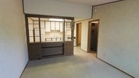 This single-floor space presents a functional and straightforward layout centered around an open-concept design. The living area is finished with neutral carpeting and light-toned walls, creating a clean and understated backdrop throughout. A standout architectural feature is the bold, dark wood open-shelving unit that serves as a partial room divider between the main living area and the kitchen. This built-in divider, framed in dark brown wood with open compartments, adds a retro design element while maintaining visual connectivity between the two spaces. The kitchen is situated just beyond the divider and is equipped with white cabinetry, a sink, and standard appliances including a range, offering a practical and organized cooking space. Overhead lighting in the kitchen provides direct illumination to the work area. A doorway visible from the main living space leads to additional rooms, suggesting a connected corridor or hallway accessing further private areas of the home. Crown molding details are present along the upper walls, adding a subtle finishing touch to the interior. The overall ambiance is understated and move-in ready, with a layout that offers clear spatial definition through built-in design elements rather than full walls.