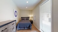 This well-appointed single-floor unit offers a cohesive and functional layout across several distinct areas, all unified by warm-toned hardwood flooring and neutral wall colors that create a bright, welcoming atmosphere.

The bedroom is a cozy retreat, featuring recessed ceiling lighting, a large window fitted with blinds for adjustable natural light, and ample space for a full-size bed and dresser. A bedside lamp adds a soft, ambient glow to the room.

The living area is comfortably furnished with a sectional sofa, a teal accent chair, and a round wooden coffee table. Large floor-to-ceiling windows flood the space with natural light and offer views of lush greenery outside. Coastal-themed artwork adorns the walls, complementing the relaxed aesthetic of the room.

The kitchen is efficiently designed with a clean, modern aesthetic. White cabinetry, a two-burner gas cooktop, an over-the-range microwave, a stainless steel refrigerator, and a dishwasher are all neatly arranged along a single wall, making excellent use of the available space.

The bathroom is clean and functional, featuring a combination shower and bathtub, a white vanity with a single sink, a large mirror, and the same warm hardwood flooring found throughout the unit, tying the space together seamlessly.