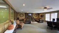 This single-story space centers around a generously sized living area that exudes a traditional, comfortable ambiance. The focal point of the room is a full brick fireplace flanked by a wall-mounted television, creating a classic yet functional gathering area. The fireplace surround extends from floor to ceiling, adding architectural character and visual weight to the space.

The seating arrangement includes dark brown leather sofas and patterned upholstered armchairs positioned on a neutral area rug, offering a cohesive and inviting layout. Decorative medallion-style wall art in earthy tones is symmetrically placed on the warm taupe walls, enhancing the traditional aesthetic.

Recessed lighting throughout the ceiling provides even illumination, complemented by a ceiling fan that adds both airflow and ambient light. The neutral-toned carpet extends across the open floor plan, contributing to the room's warm feel. A window along one wall allows natural light to enter, brightening the space. The textured ceiling and understated color palette tie all elements together, resulting in a cohesive, welcoming environment.