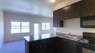 This single-floor space showcases an open-concept layout that seamlessly connects the kitchen and living area. The kitchen is a standout feature, equipped with rich, dark espresso-toned cabinetry that extends both above and below the countertops, providing generous storage throughout. A peninsula with matching dark cabinetry serves as a natural divider between the kitchen and living space, offering additional prep and storage space while maintaining an open feel. The countertops complement the cabinetry with a clean, dark finish, and a stainless steel sink is positioned along the perimeter wall, accented by a subtle mosaic tile backsplash that adds a touch of visual interest. Upper cabinets extend toward the ceiling, maximizing vertical storage. The living area benefits from multiple windows that invite an abundance of natural light, creating a bright and welcoming atmosphere. A sliding glass door provides access to an outdoor space, further enhancing the connection between interior and exterior. The textured ceiling and neutral-toned walls keep the aesthetic clean and understated, allowing the dark kitchen elements to serve as the focal point throughout the open space.