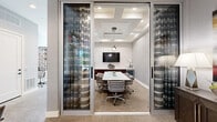 Located at 3900 W Northwest Highway in Dallas, TX, this sophisticated business lounge offers a well-appointed environment designed for professional productivity. The space is accessed through floor-to-ceiling glass walls and doors framed in sleek aluminum, providing excellent visibility and an open, modern aesthetic enhanced by bold striped drapery panels.

The centerpiece of the space is a private conference room featuring a coffered ceiling with recessed lighting and a striking sculptural pendant fixture. A large white conference table is surrounded by upholstered rolling chairs, while a wall-mounted flat-screen TV and rich dark wood credenza with built-in beverage cooler offer both functionality and refined style. Abstract artwork adorns the walls, adding a creative touch to the professional setting.

Adjacent to the conference room, a dedicated workstation area provides a long built-in desk with a computer and printer, complemented by decorative wood shelving and unique circular wall sconces. The lounge seating area features a comfortable sofa and accent chairs arranged around a coffee table, creating a relaxed setting for informal meetings or waiting. Neutral carpet flooring, textured wall coverings, and consistent recessed lighting throughout tie the entire space together with a cohesive, contemporary design.